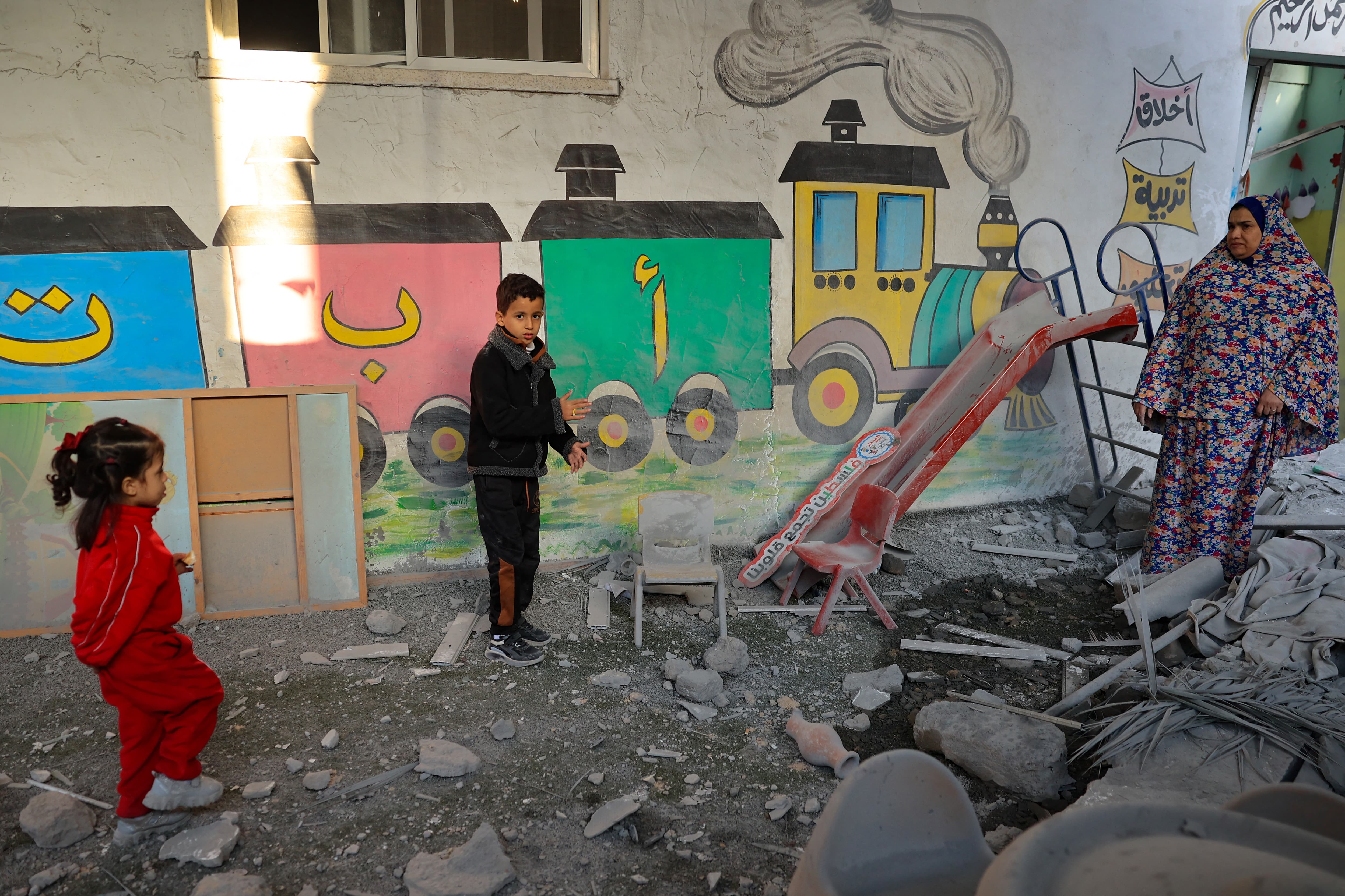 A Palestinian woman and children stand amid the rubble at a kindergarten hit by Israeli air strikes in Rafah, in the southern Gaza Strip, on 9 December 2023 (Said Khatib/AFP)