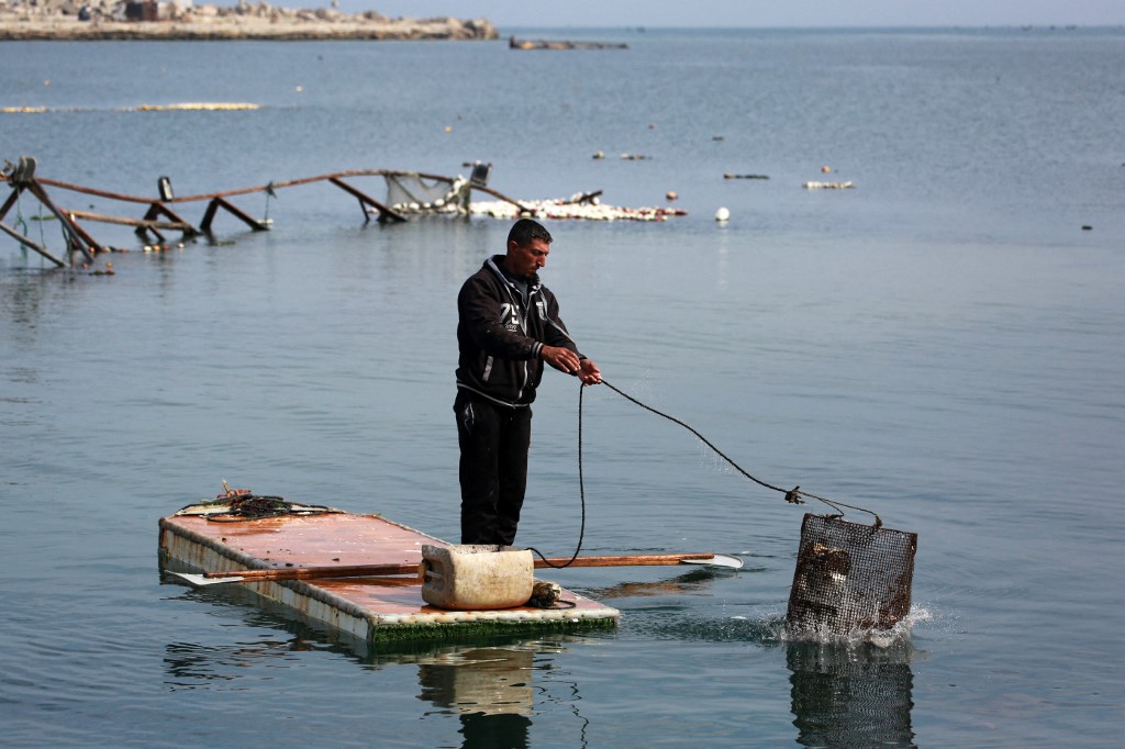 A Palestinian man stands on the door of a refrigerator that he uses as a makeshift rowing boat, as he throws his fishing basket into the sea at the port of Gaza City on March 9, 2025. (Bashar Taleb/AFP)