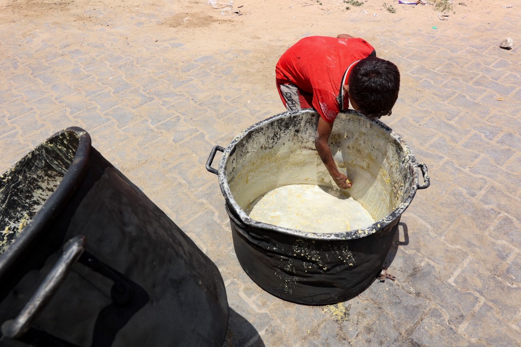 A Palestinian boy scrapes off bits of lentil soup remaining in a cooking pot, in front of a food distribution point at a displacement camp near Gaza City's port on May 22, 2025.