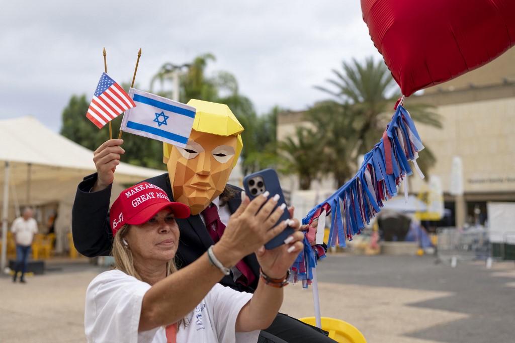 A person wearing a mask depicting US President Donald Trump holds US and Israeli flags after the hostage deal declared by Trump, at Hostage Square in Tel Aviv on October 9, 2025.