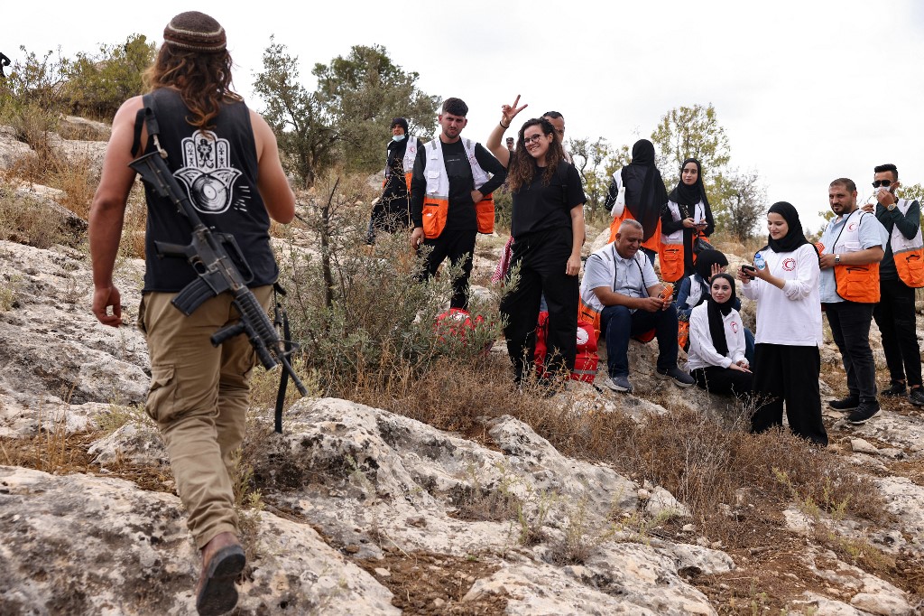 An Israeli settler walks uphill past an activist and Red Crescent rescue workers in the village of Idna, north of the occupied-West Bank city of Hebron, during the olive harvesting season, on October 12, 2025