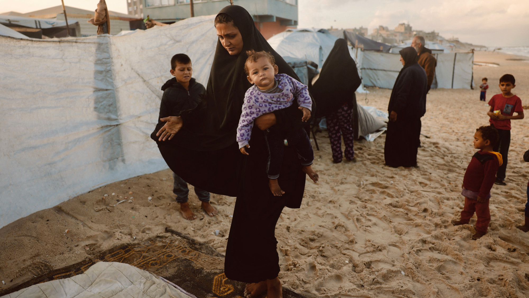 A Palestinian woman carries her child as they shelter from the rain in a makeshift camp in Deir al-Balah, central Gaza (AFP/Bashar Taleb).