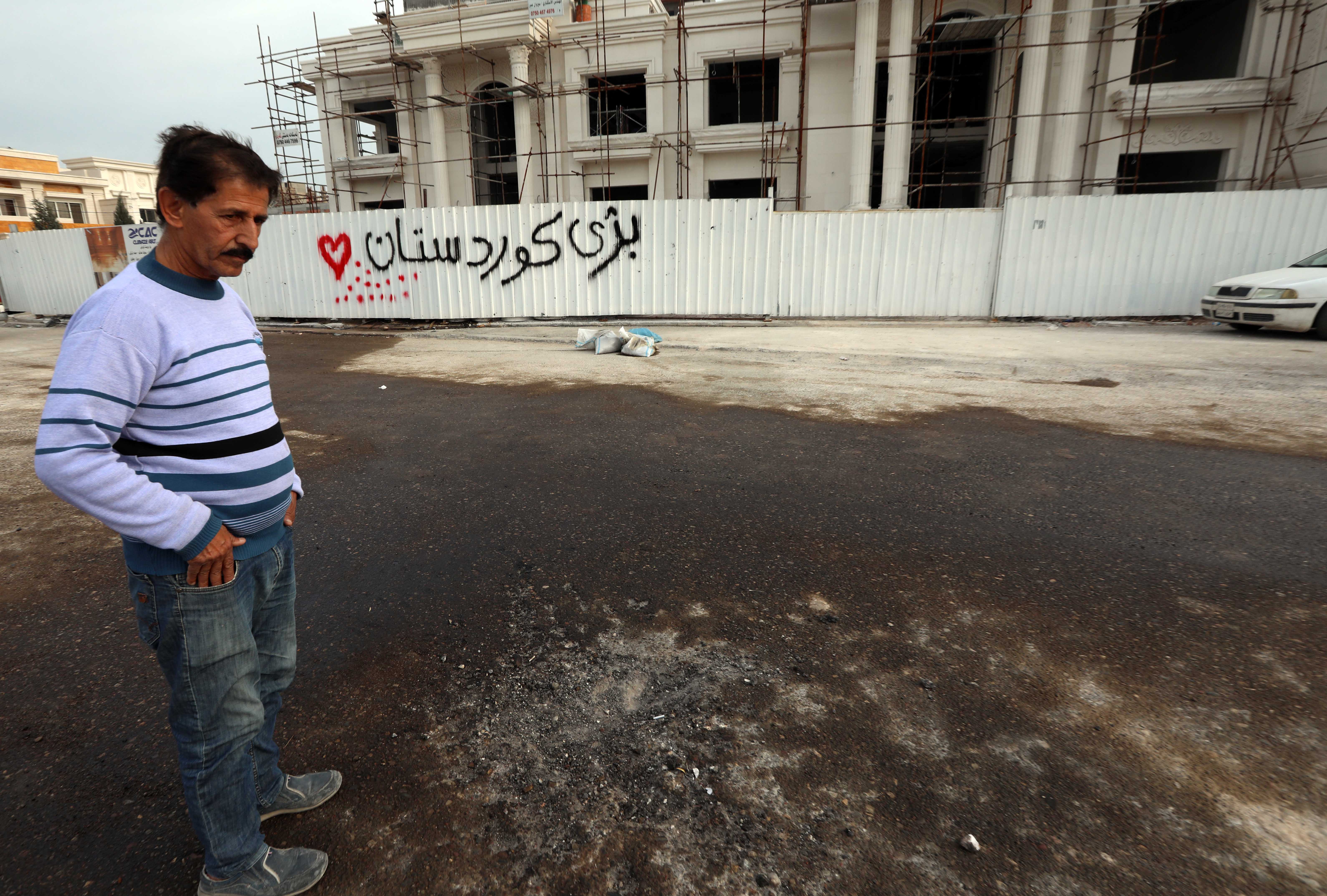 A man stands near a fence with graffiti reading in Kurdish 