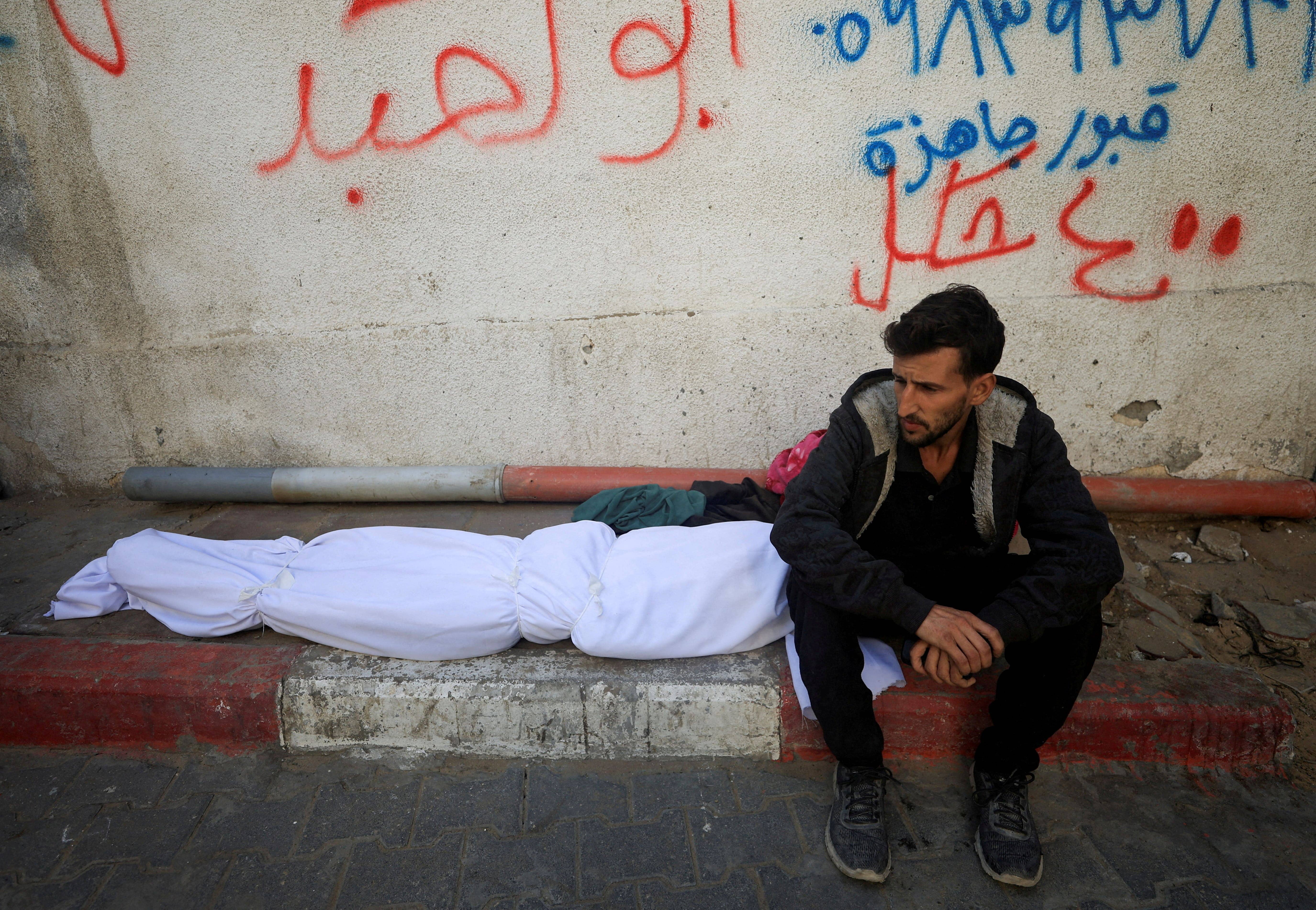 A mourner sits next to the body of a Palestinian who, according to medics, was killed in overnight Israeli strikes, at Al-Shifa Hospital in Gaza City, 20 November 2025. (Reuters)