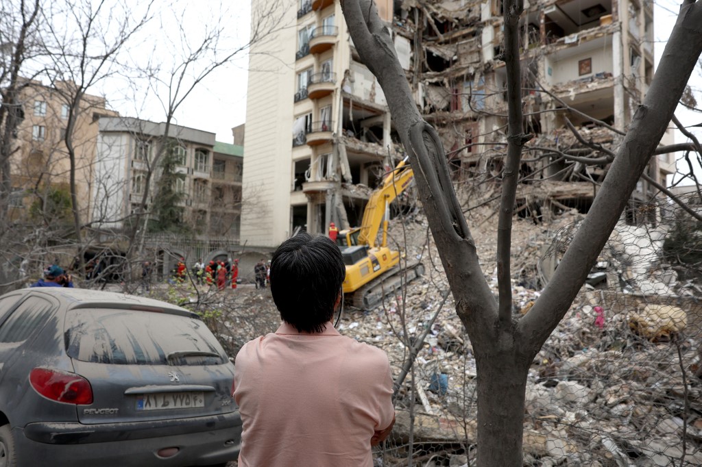 A resident looks on as Iranian firefighters with the help of an excavator clear rubble from a destroyed residential building in northern Tehran on 23 March, 2026. (AFP)