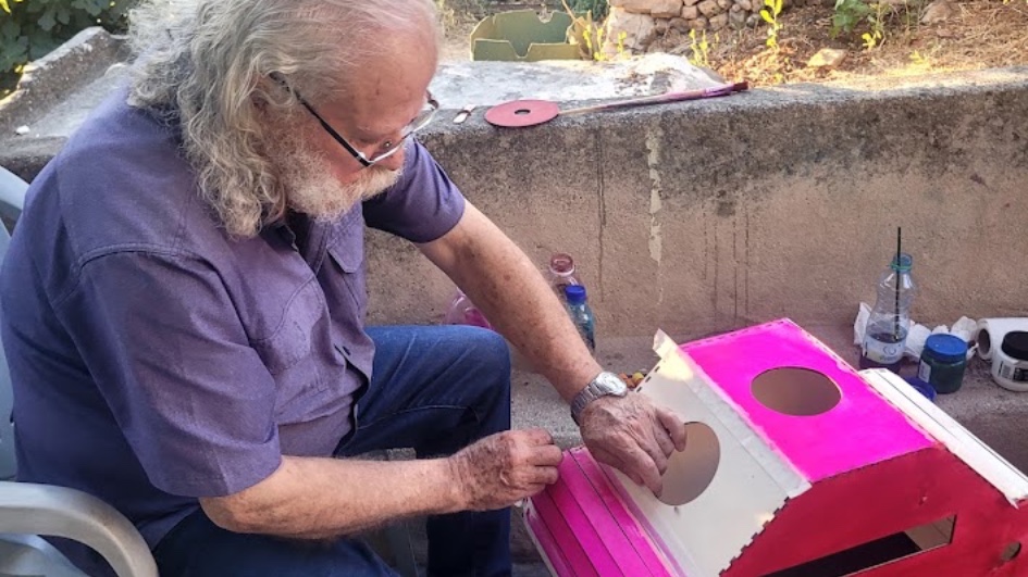An elderly man with long white hair paints a wooden Wonderbox bright pink