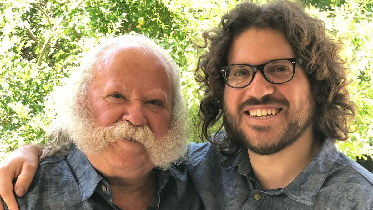 An older man with a white beard stands with his arm around his adult son, both smiling