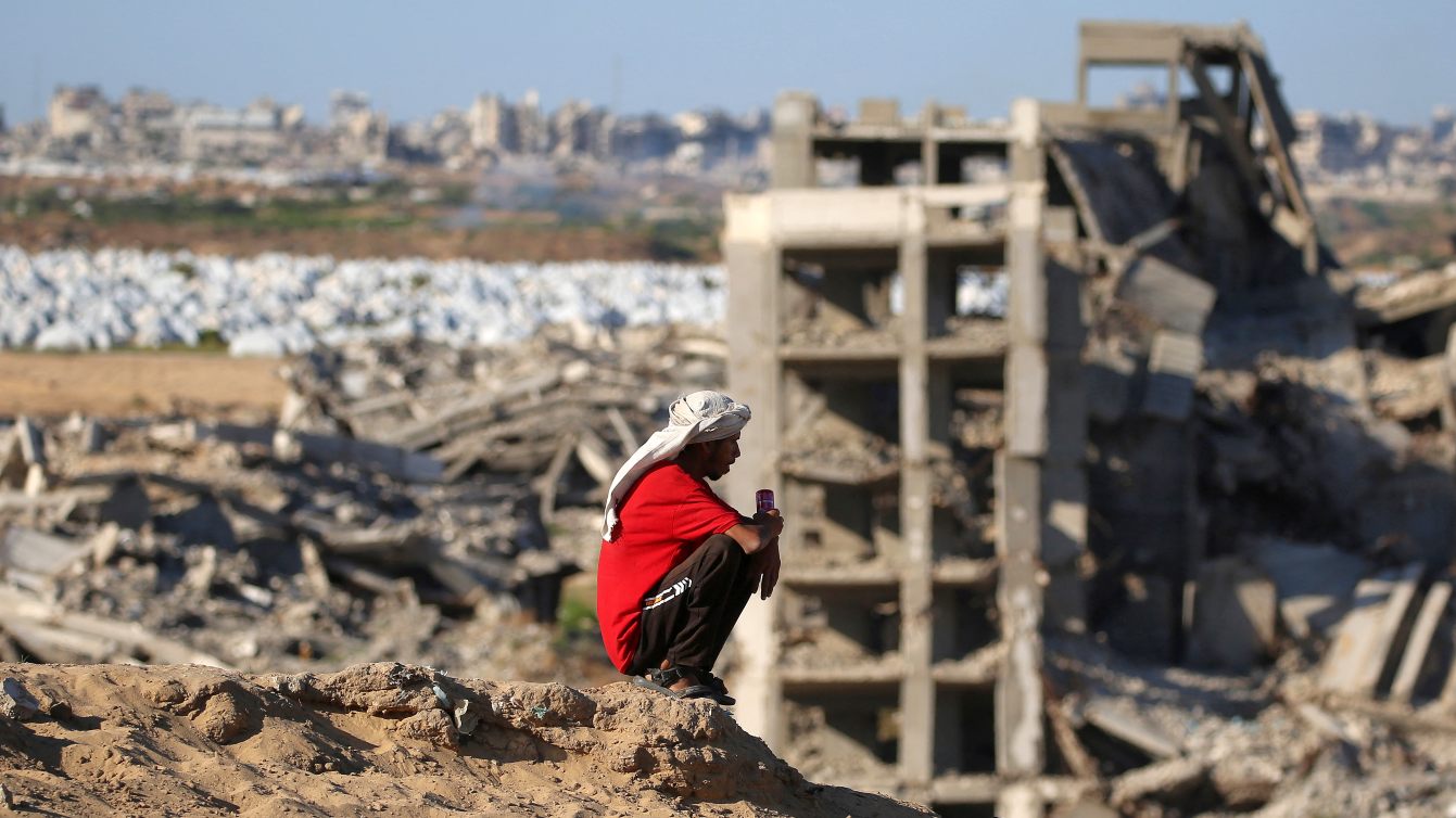 Man sits atop a hill overlooking destroyed and heavily damaged buildings in Nuseirat refugee camp in central Gaza, 19 June 2026 (Eyad Baba/AFP)
