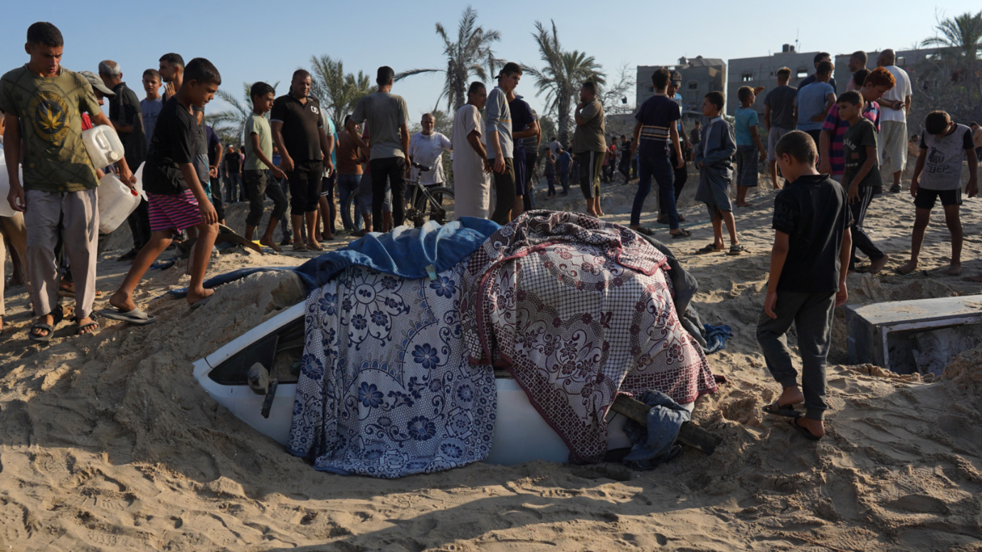 Palestinians stand next to a car covered in sand at the site of Israeli strikes on a refugee camp in al-Mawasi, southern Gaza (Bashar Taleb/AFP)