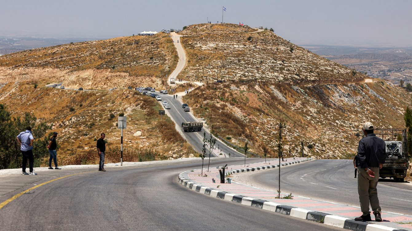 Palestinian men watch from a roadside as Israeli settlers and Finance Minister Bezalel Smotrich attend a settlement ceremony near Hebron in the occupied West Bank, 16 June 2026 (Hazem Bader/AFP)