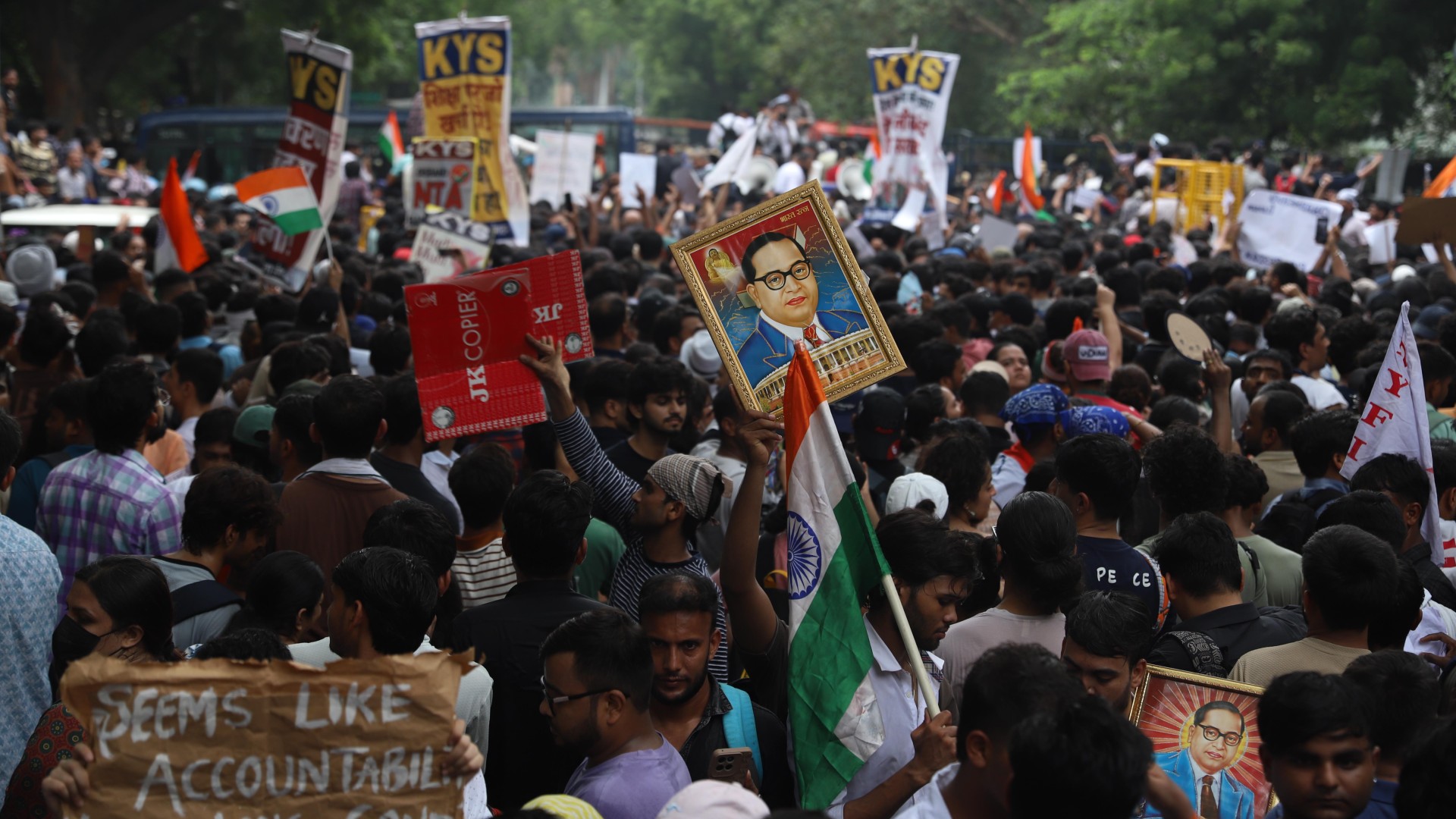 A large crowd of protesters march through a tree-lined street, waving Indian flags and holding placards demanding accountability and the resignation of the Indian education minister