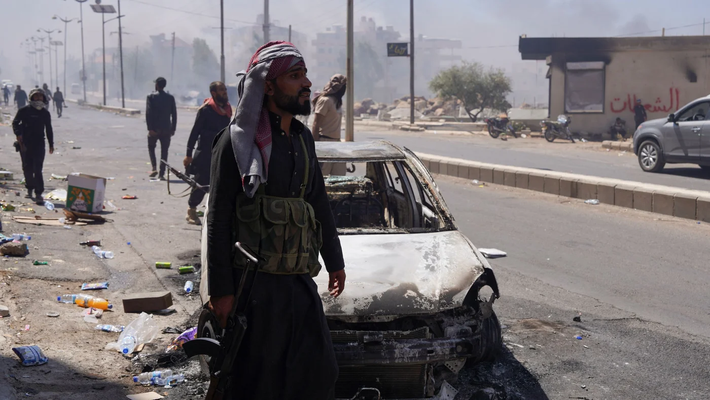 A Bedouin fighter stands near a damaged car in Sweida after the Syrian presidency announced a ceasefire following days of clashes with Druze factions, on 19 July 2025 (Reuters/Karam al-Masri)