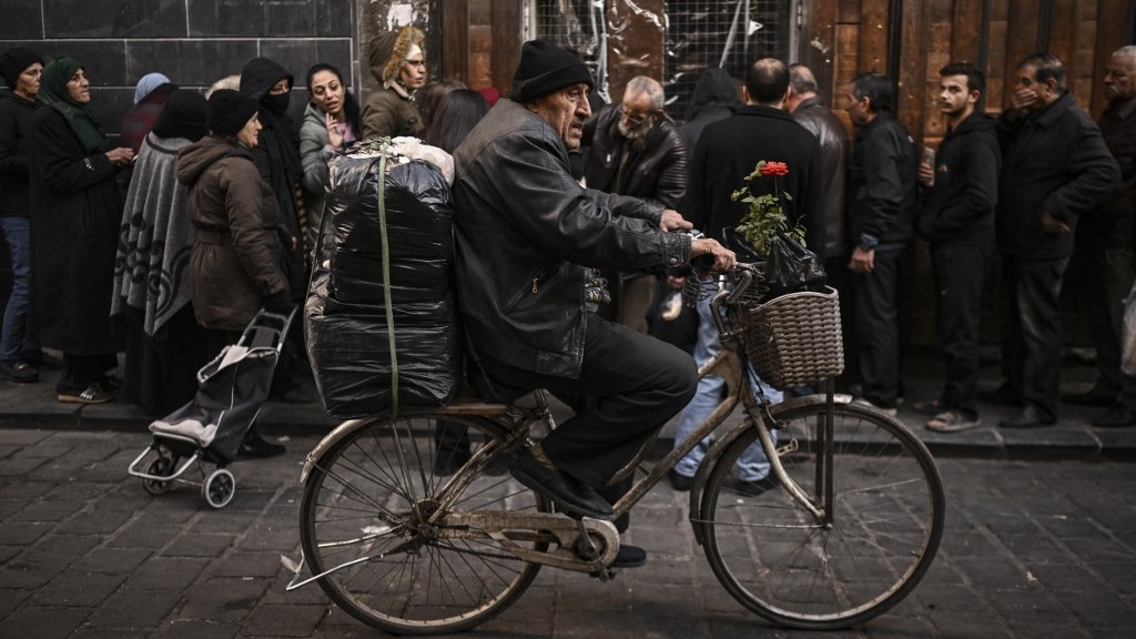 A man rides his bicycle past people lining up to buy bread in the Syrian capital Damascus on December 21, 2024. Islamist-led rebels took Damascus in a lightning offensive on December 8, ousting president Bashar al-Assad and ending five decades of Baath rule in Syria.