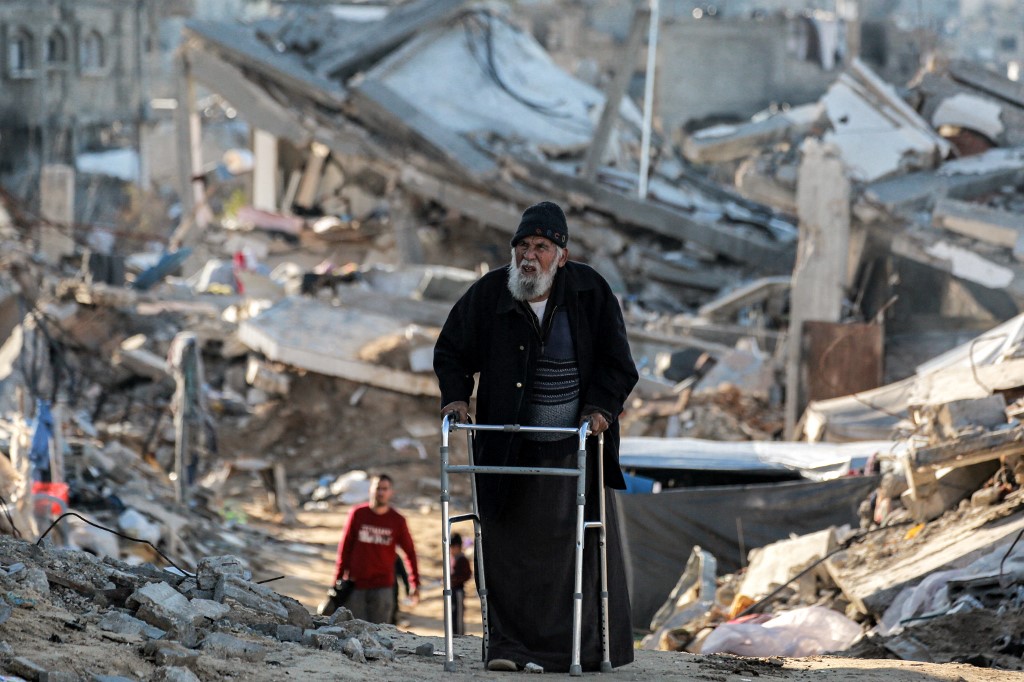 An elderly man moves with a walker past rubble along a broken road as people displaced by conflict from Beit Lahia arrive in Gaza City on March 22, 2025. 