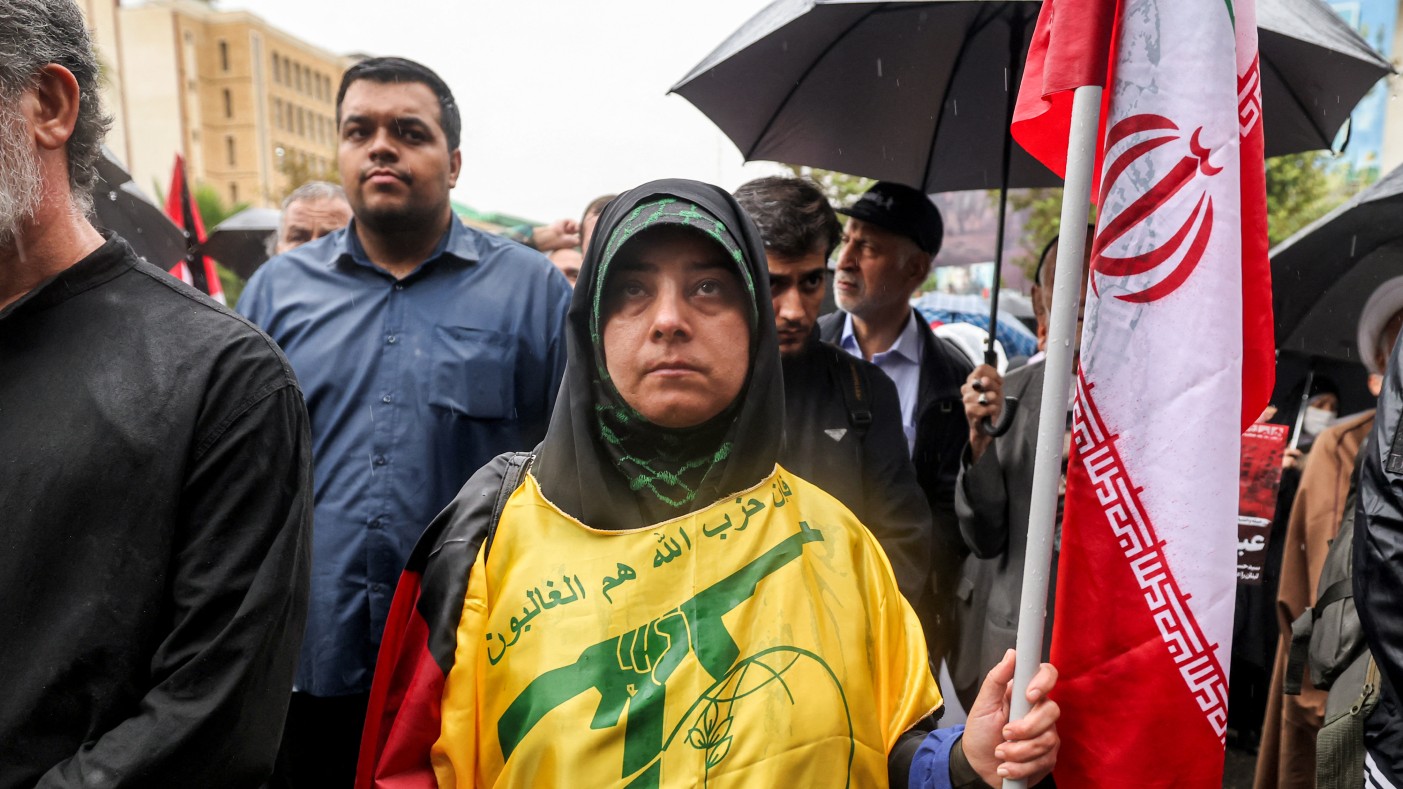 A woman stands holding an Iranian flag and draped in the yellow flag of the Hezbollah