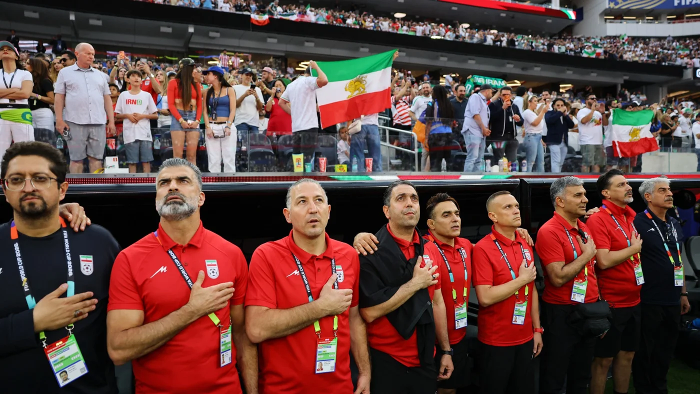 Members of the Iran coaching team during the national anthems before the New Zealand match as fans display pre-Iranian Revolution 'Lion and Sun' flags behind them, 15 June (Reuters/Daniel Cole)