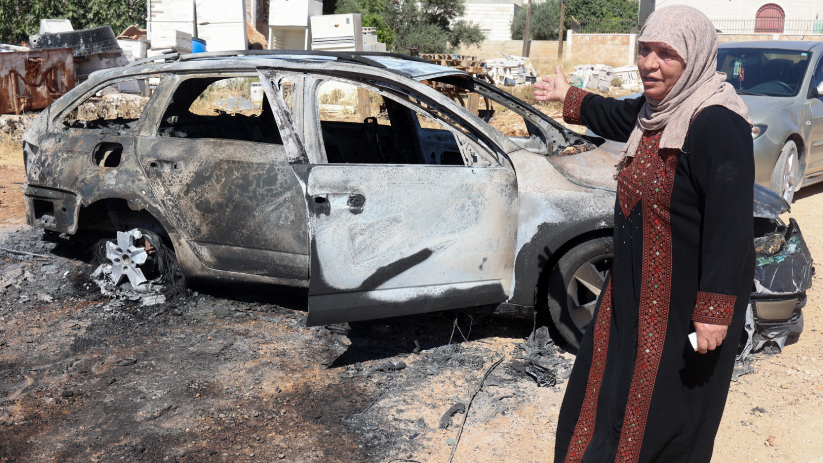 A Palestinian woman gestures in front of a burnt car after an Israeli settlers attack the previous day in Kafr Malik in the Israeli occupied West Bank, on 26 June 2025 (AFP)