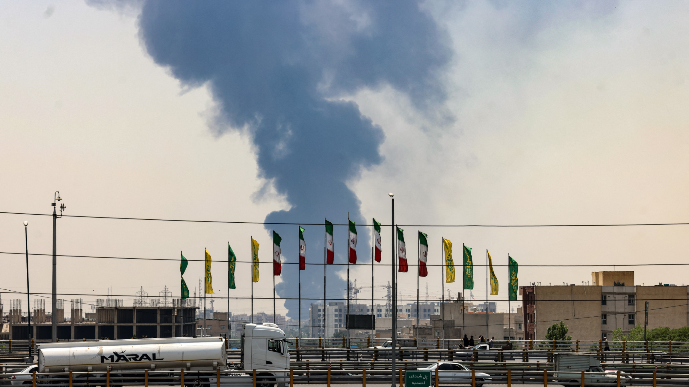 Flags flutter along a bridge as a plume of heavy smoke and fire rise from an oil refinery in southern Tehran, after it was hit in an overnight Israeli strike, on 15 June 2025 (AFP)