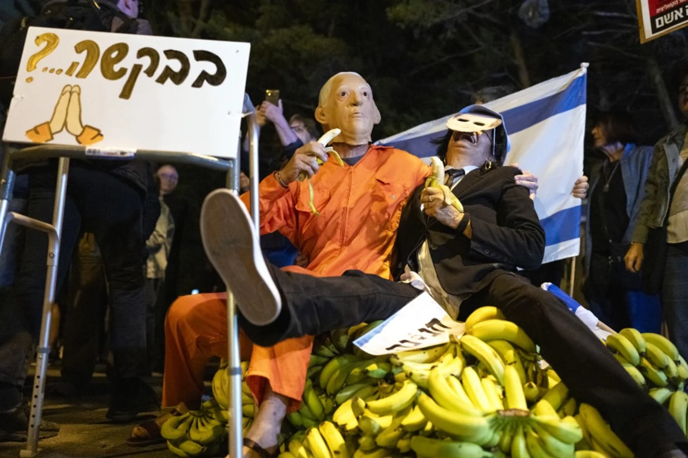 A demonstrator wearing a mask depicting Benjamin Netanyahu sits on a pile of bananas with a man pretending to be Isaac Herzog in Tel Aviv on 30 November 2025 (Maya Levin/AFP)