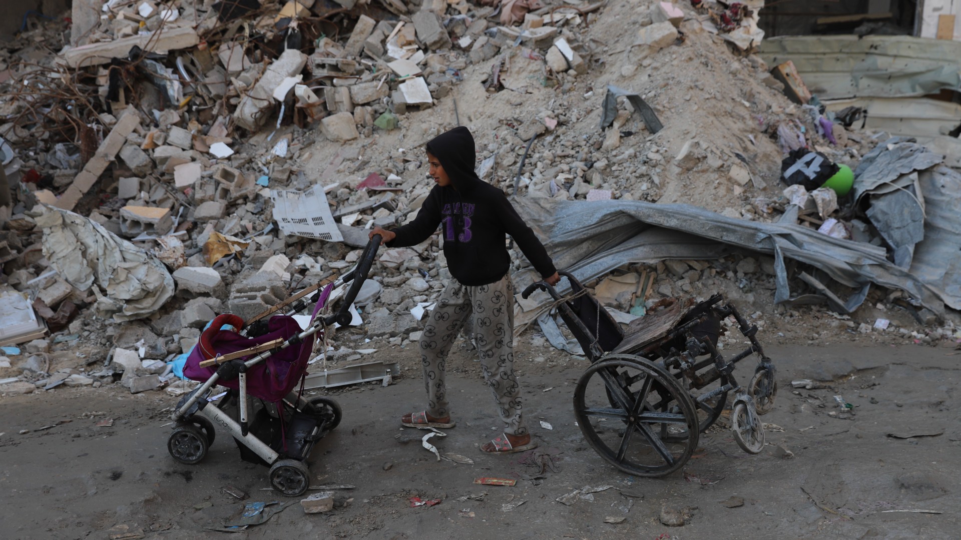 A Palestinian child pushes a pram and a wheelchair through a destroyed road in Gaza City on 2 February 2025 (Mohammed al-Hajjar/MEE)