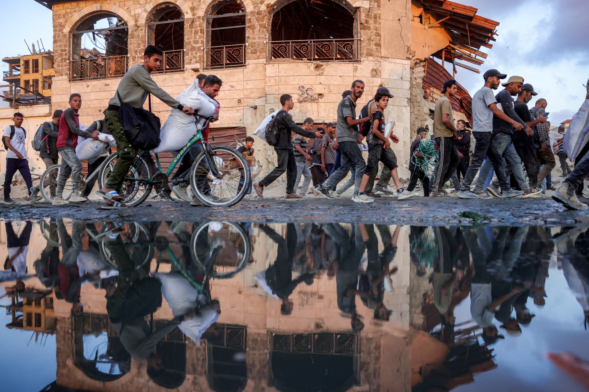 People carrying sacks of flour walk past a water puddle along al-Rashid street in western Jabalia on 17 June 2025 (AFP/Bashar Taleb)