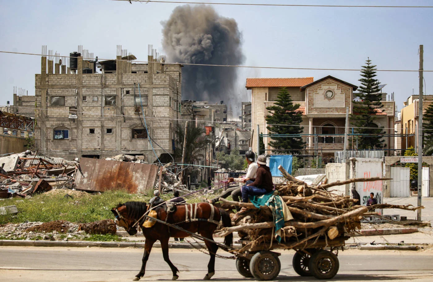 Men riding a horse-drawn cart carrying chopped wood watch as a smoke plume erupts from an Israeli air strike in central Gaza on 25 March 2025 (AFP)