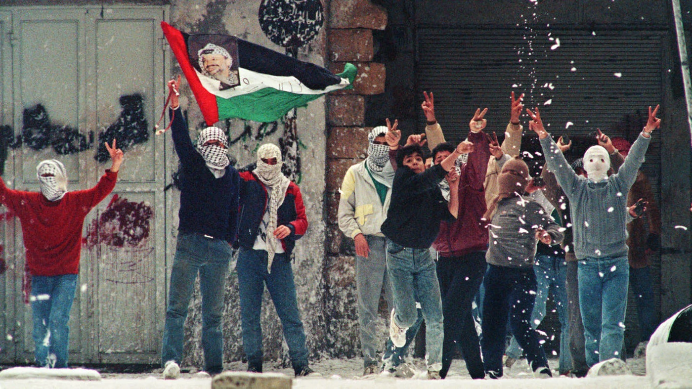 Palestinian demonstrators hold a Palestinian flag with a portrait of PLO leader Yasser Arafat as foam thrown by Israeli soldiers drops from a roof in Nablus, West Bank, in January 1988 (AFP)