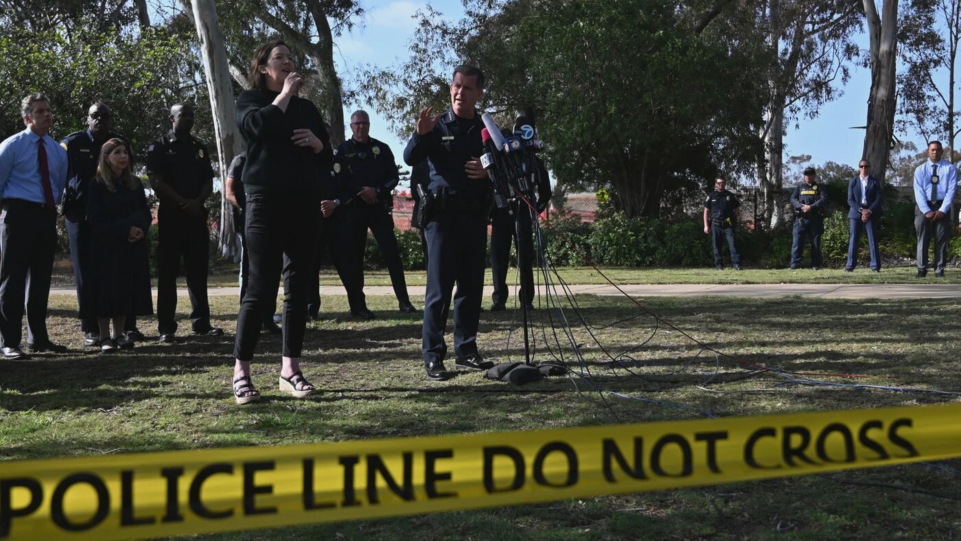 Police officers and officials stand behind a podium of microphones at an outdoor press briefing, with yellow "police line do not cross" tape stretched across the foreground