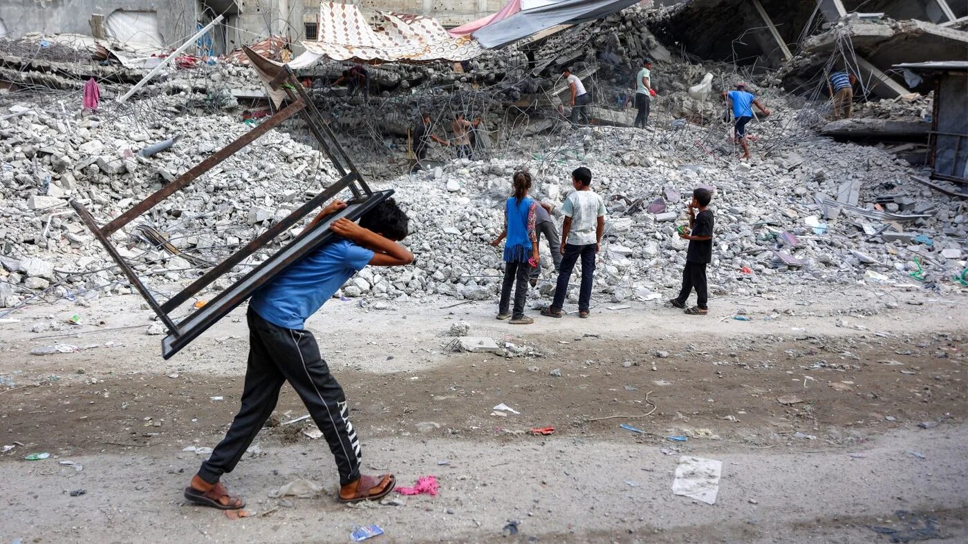 A boy carries a table past workers salvaging rubble from a building destroyed during the war in Gaza City, 16 August 2026 (Omar Al-Qattaa/AFP)