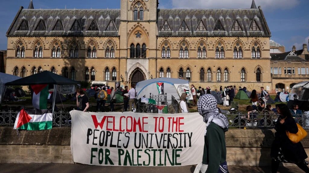 A passerby with the head covered with a keffiyeh walks past a banner reading "Welcome to the people's university for Palestine" at Oxford University, in Oxford, eastern England on May 7, 2024