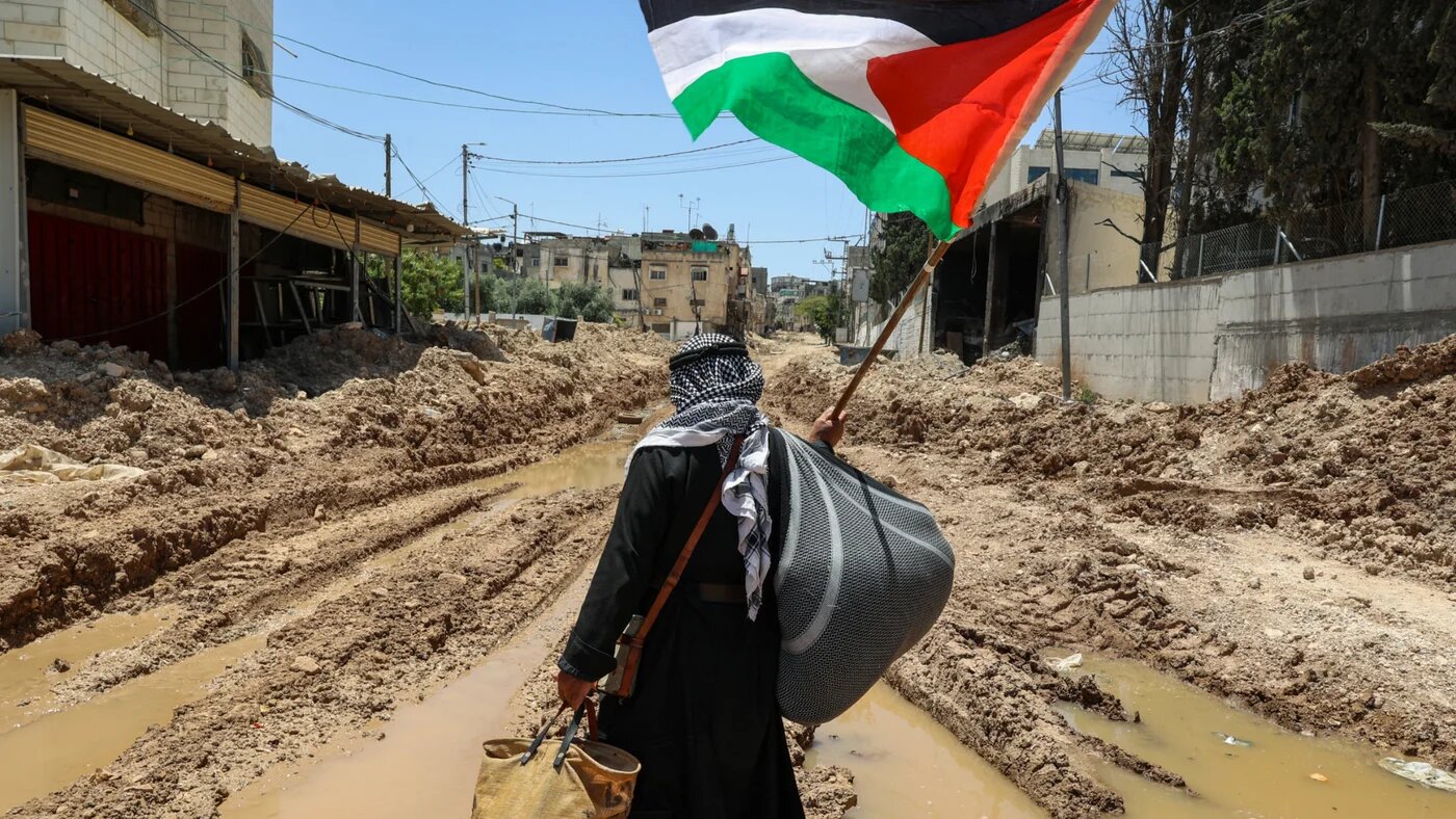 A Palestinian man lifts a national flag as he walks on a road destroyed by Israeli forces in the Tulkarm refugee camp in the occupied West Bank on 19 May 2025 (AFP/Zain Jaafar)