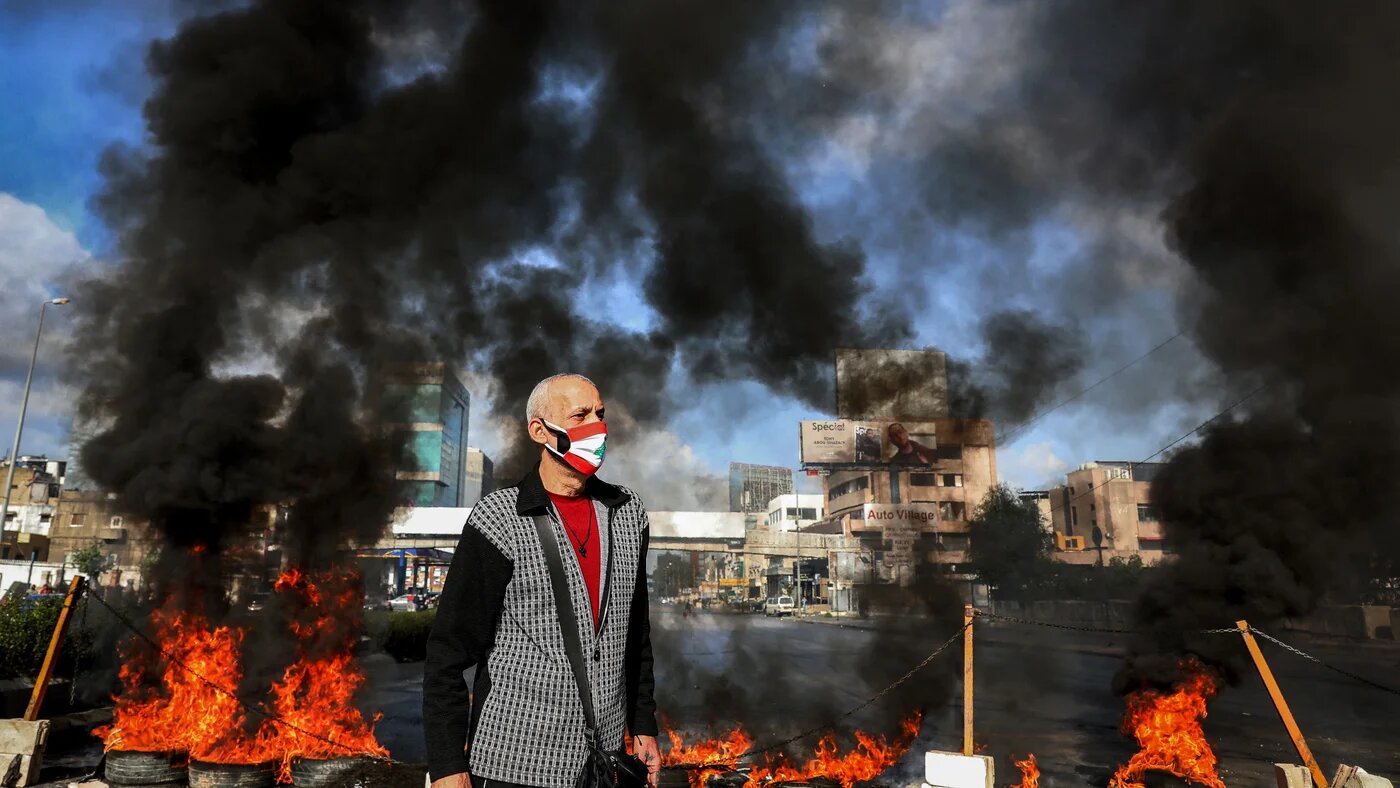 A man in mask a depicting the Lebanese flag stands next to flaming tires at a make-shift roadblock set-up by anti-government demonstrators in  Dora on the northern outskirts of Lebanon's capital Beirut on 8 March 8 2021 (Anwar Amro/AFP)