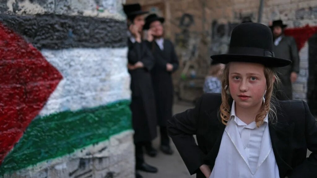 A young member of the Neturei Karta, an international organisation of anti-Zionist Orthodox Jews, poses for a picture next to a mural of a Palestinian flag during a protest against the State of Israel in  Jerusalem on 22 April 2026 (Ahamad Gharabli/AFP)