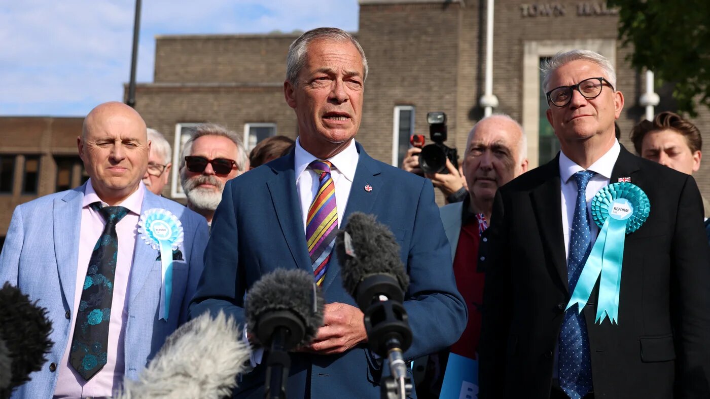 Reform UK leader Nigel Farage is pictured in a blue suit and a multi-coloured tie with microphones pointed at him as he speaks to the press