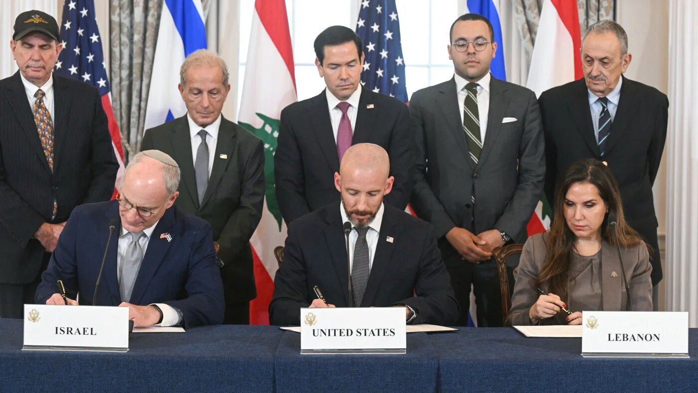 Three officials sign documents at a long table beneath Israeli, US and Lebanese flags