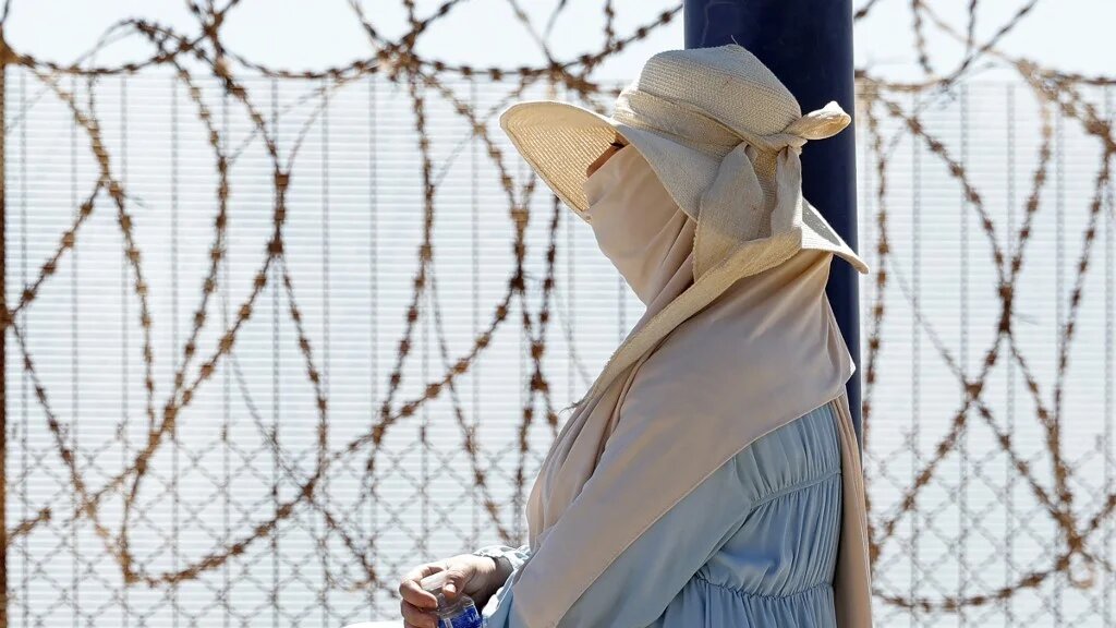 A woman sits by a barbed-wire fence as relatives await the return of people from Spain's North African enclave of Ceuta at the Bab Sebta border crossing near Fnideq, Morocco, 2 August 2026 (Abdel Majid Bziouat/AFP)