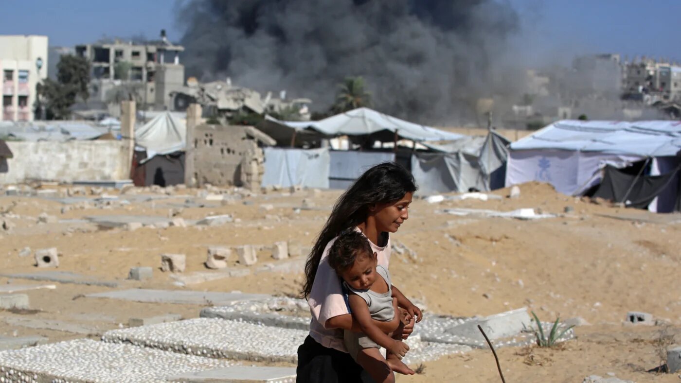 A Palestinian girl carries a child against the backdrop of smoke rising from a house that was targeted by an Israeli strike in Khan Yunis, southern Gaza Strip on 9 September 2026 (AFP/Bashar Taleb)