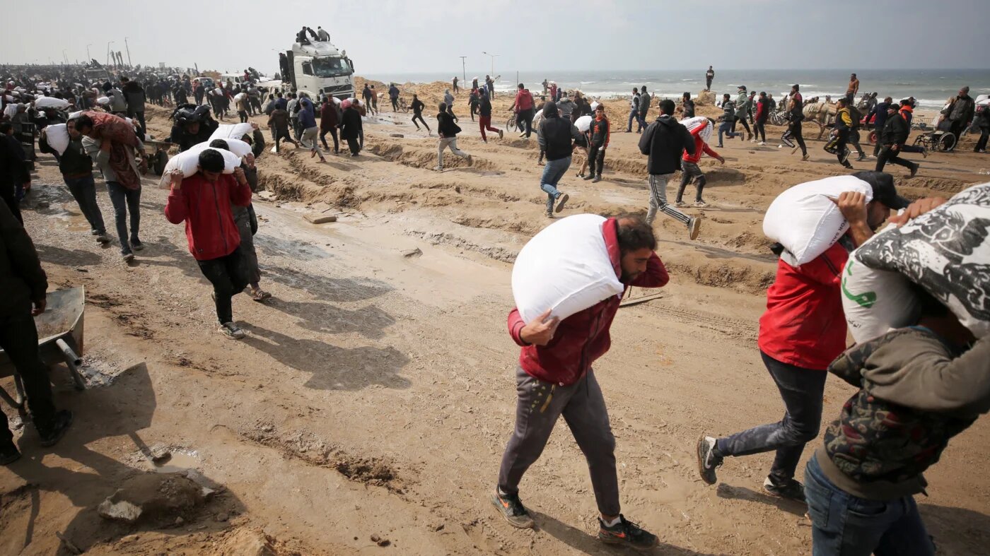Palestinians carry bags of flour they grabbed from an aid truck near an Israeli checkpoint, as Gaza residents face crisis levels of hunger in Gaza City, 19 February 2024 (Reuters/Kosay Al Nemer)