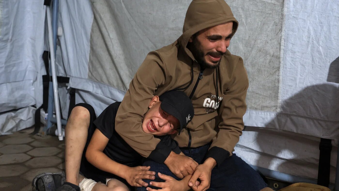 A man and a child mourn by the covered body of a Palestinian killed in an Israeli air strike, according to medics, at Al-Ahli Arab Hospital in Gaza City 19 November 2025 (Reuters/Dawoud Abu Alkas)