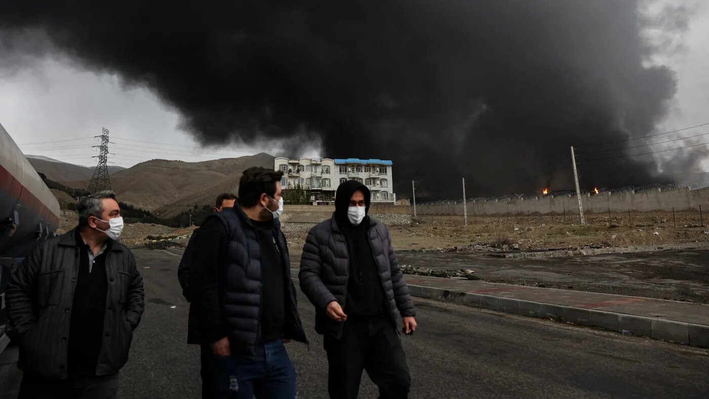 People walk as smoke rises in the background after a reported strike on the Shahran fuel tanks in Tehran, Iran, on 8 March 2026 (WANA/Majid Asgaripour via Reuters)