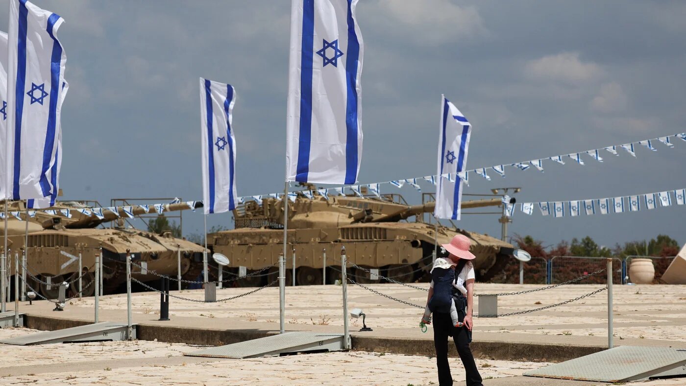A woman carries a child near Israeli flags and tanks during Memorial Day commemorations in Latrun, the eve of Israel's 78th Independence Day, 21 April 2026 (Florion Goga/Reuters)