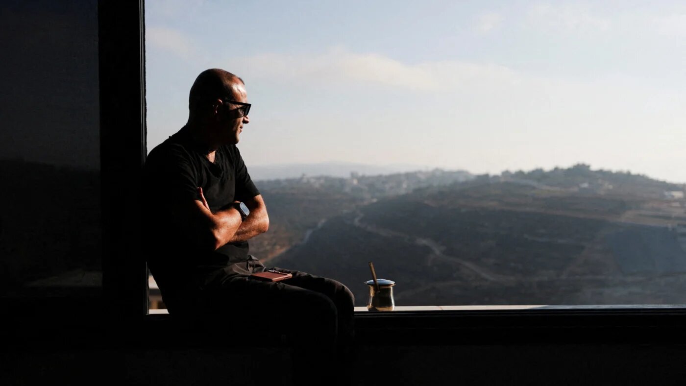 Palestinian-American Loui Ridi looks out the window of his home, which has been encircled by settlers for two weeks in Qusra, occupied West Bank, 18 August 2026 (Ammar Awad/Reuters)