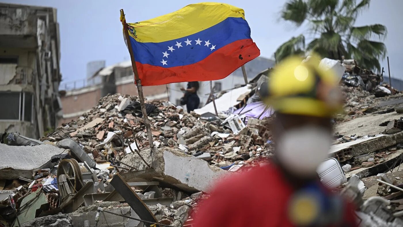 A Venezuelan flag flies over the rubble of collapsed buildings as a rescue worker stands in Caraballeda after an earthquake, 20 July 2026 (Federico Parra/AFP)
