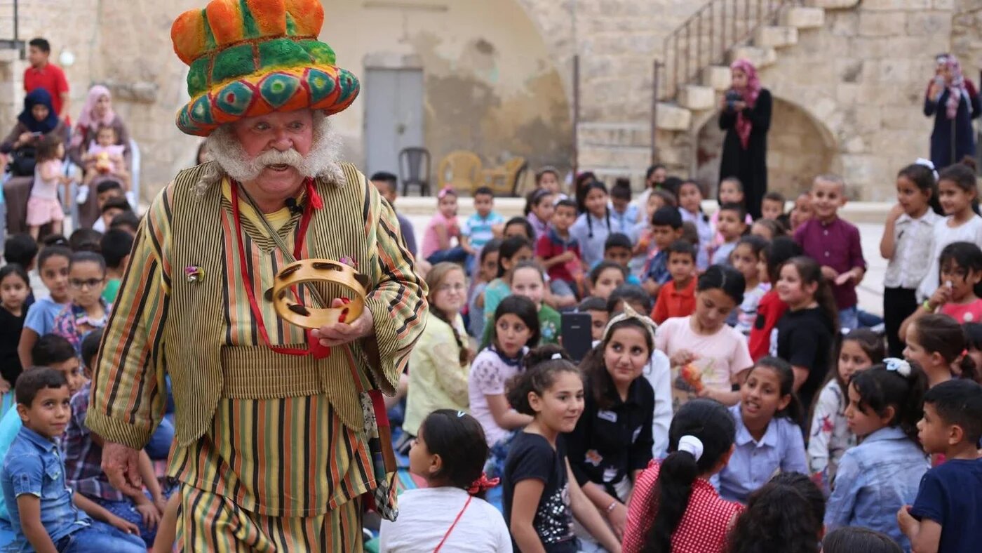 Adel Tartir, in a striped costume and tall colourful hat, plays a tambourine before a crowd of seated children