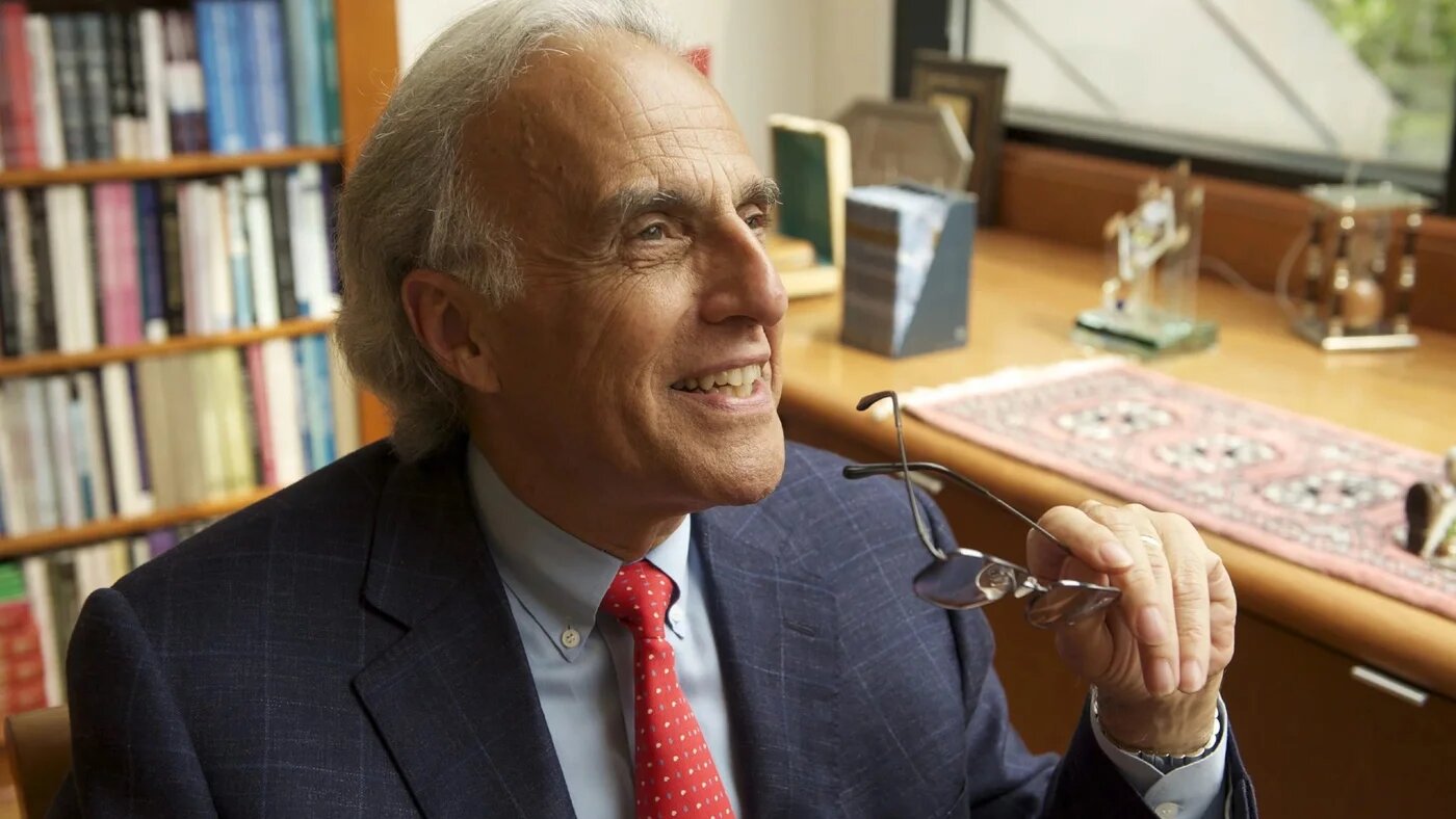 An older man in a dark suit and red tie smiles, holding his glasses, in an office lined with books