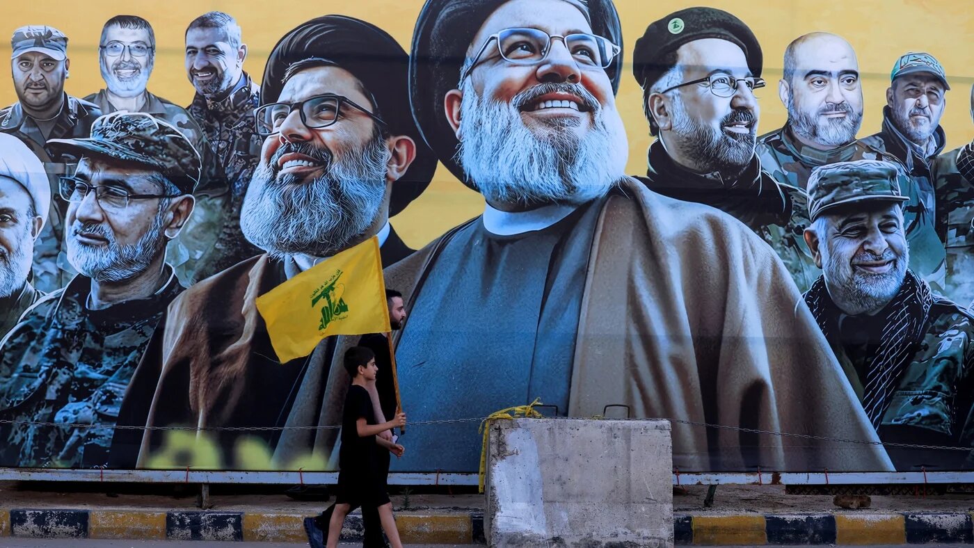 A man walks with a boy, carrying a Hezbollah flag, past a mural depicting former Hezbollah leader Hassan Nasrallah and other senior figures (Reuters//Mohamed Azakir)