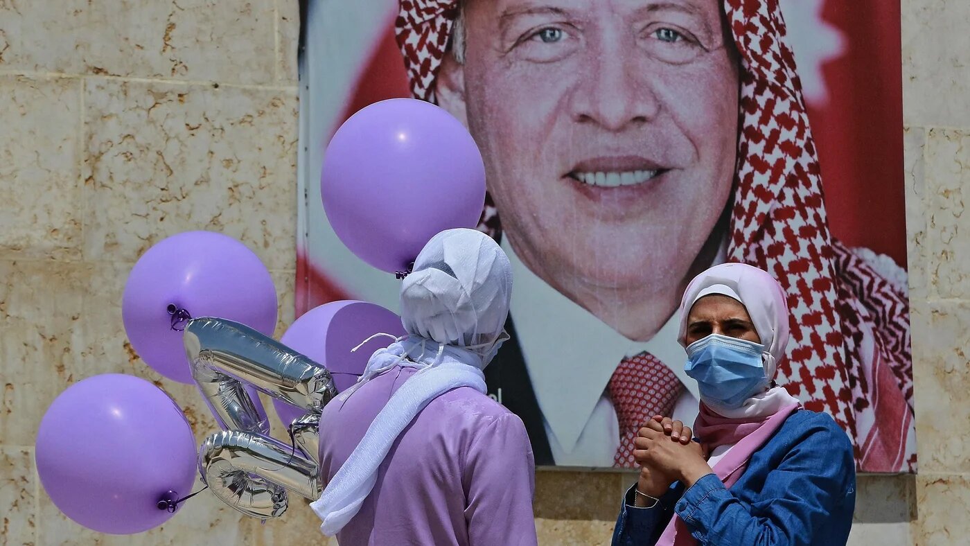 Women carrying balloons stand next to a poster of Jordan's King Abdullah in Amman