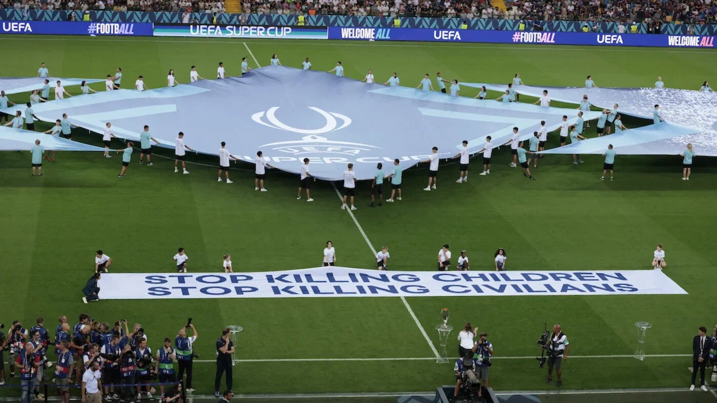 Refugee children display 'Stop killing children stop killing civilians' banner on the pitch ahead of the 2025 Uefea Super Cup final between Paris Saint-Germain and Tottenham Hotspur FC at the Friuli stadium, Udine, on 13 August 2025 (Remo Casilli/Reuters)