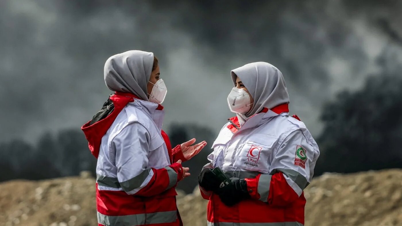 Members of Iran's Red Crescent society stand near smoke plumes from an ongoing fire following an overnight airstrike on the Shahran oil refinery in northwestern Tehran on 8 March 2026 (AFP)
