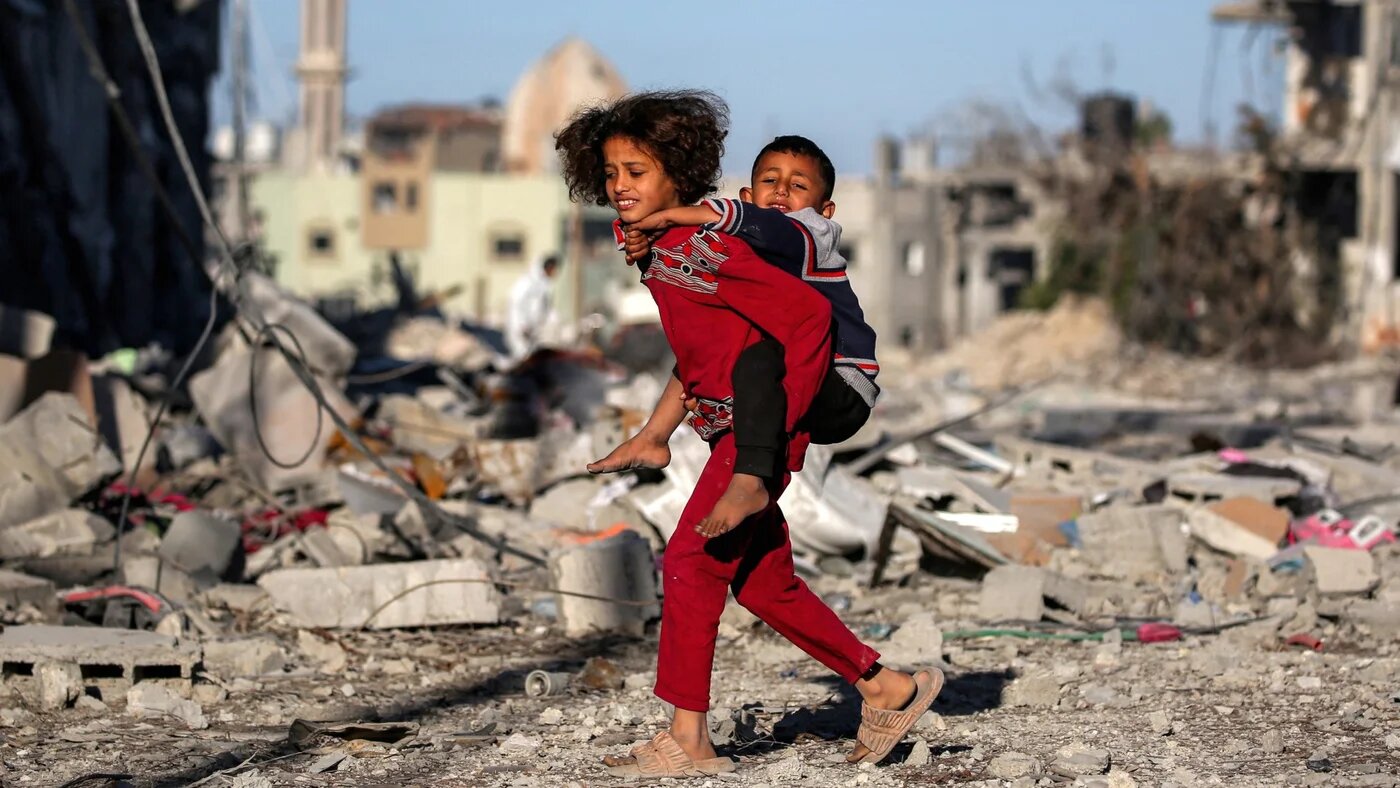 A girl carries a boy on her back as she walks past devastation unleashed by Israel on Rafah in the southern Gaza Strip on 21 January 2025 (Eyad Baba/AFP)